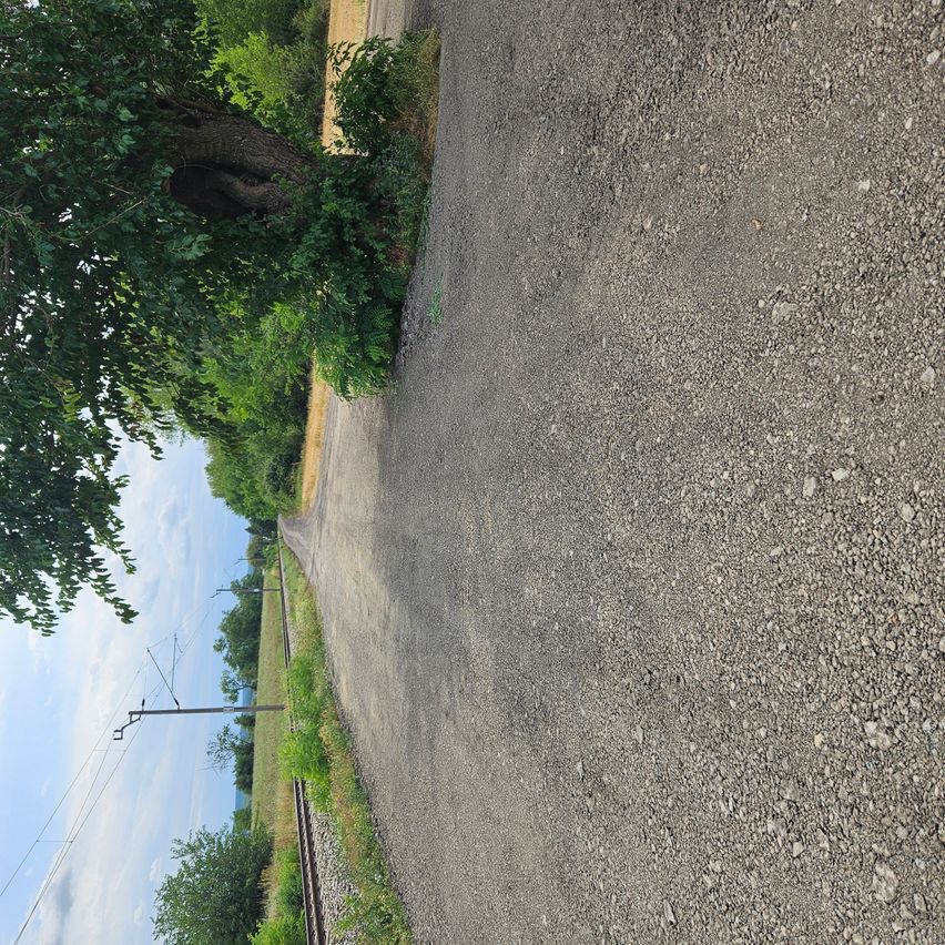 Eine Schotterstraße mit einem Baum auf der linken Seite und einer Eisenbahnstrecke auf der rechten Seite. Der Himmel ist klar mit ein paar Wolken.