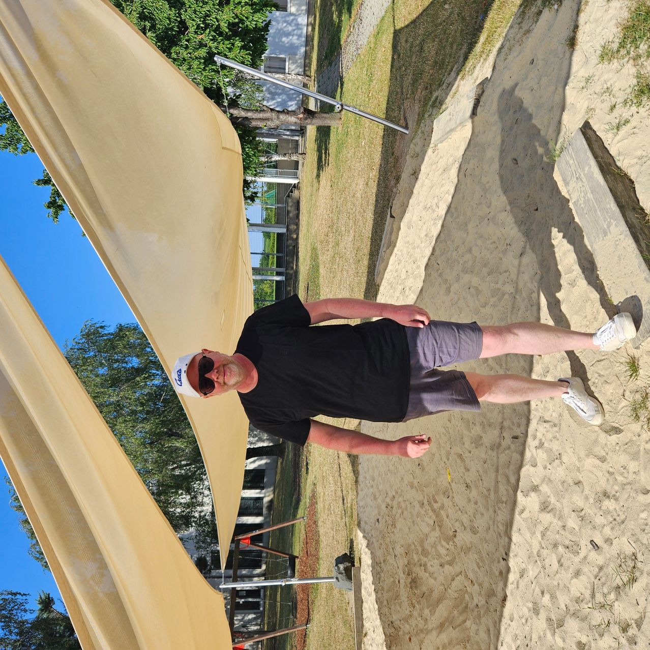 Ein Mann steht auf einem sandigen Strand unter einem beigen Vordach, trägt ein schwarzes T-Shirt, graue Shorts und weiße Turnschuhe. Er lächelt und schaut in die Kamera.