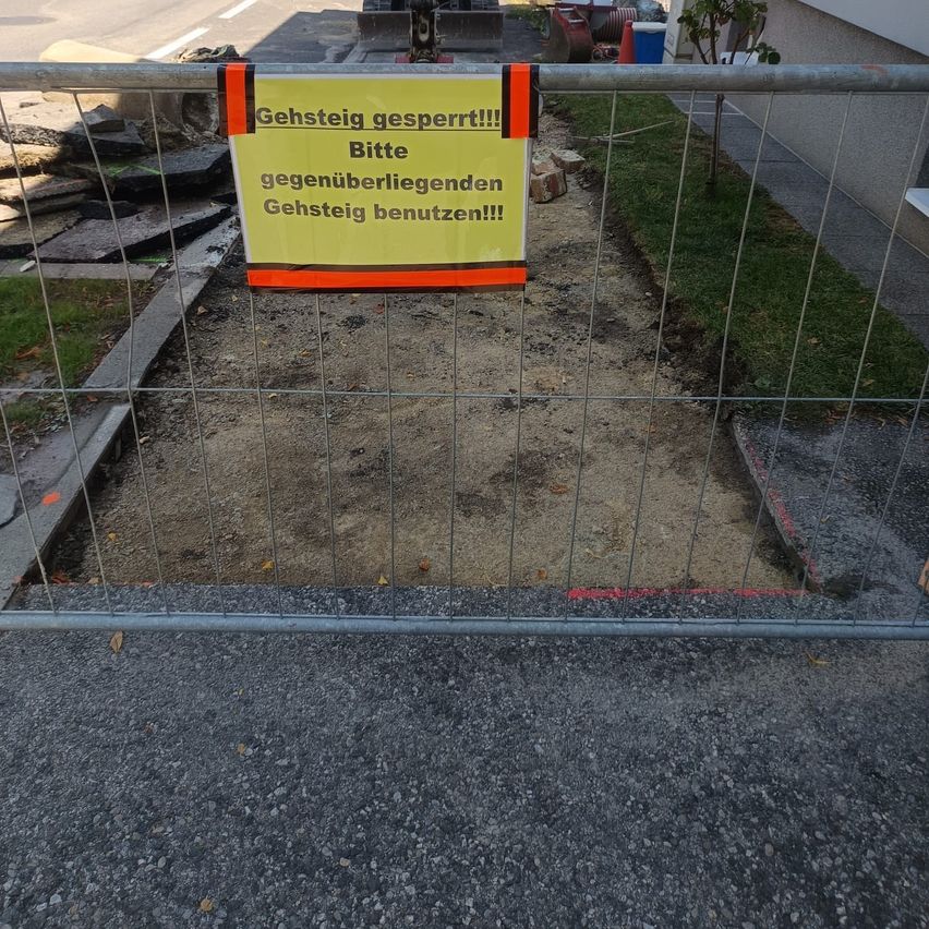 Ein Baustelle mit einem gelben Schild auf Deutsch, das 'Bitte benutzen Sie den angrenzenden Bürgersteig!' besagt, und einem Metallzaun, der den Bereich blockiert.