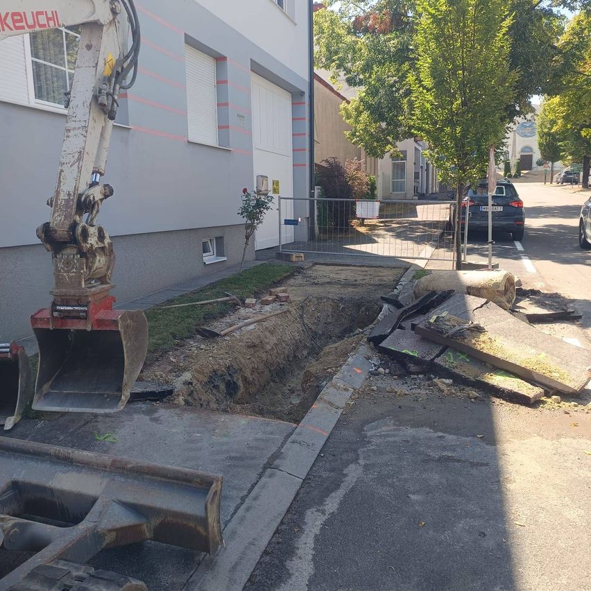Eine Baustelle mit einem Bagger, der ein Loch in den Bürgersteig gräbt. In der Nähe befindet sich ein Gebäude mit Fenstern und einem Zaun. Auf der Straße ist ein Auto geparkt.
