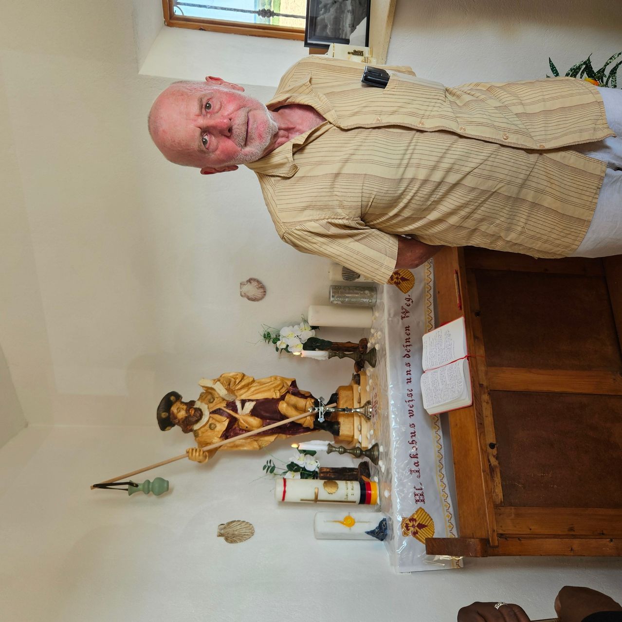 Ein älterer Mann steht in einem Raum mit einem religiösen Altar hinter ihm, der eine Statue, Kerzen und ein Buch zeigt. Er trägt ein gestreiftes Hemd und weiße Hosen.
