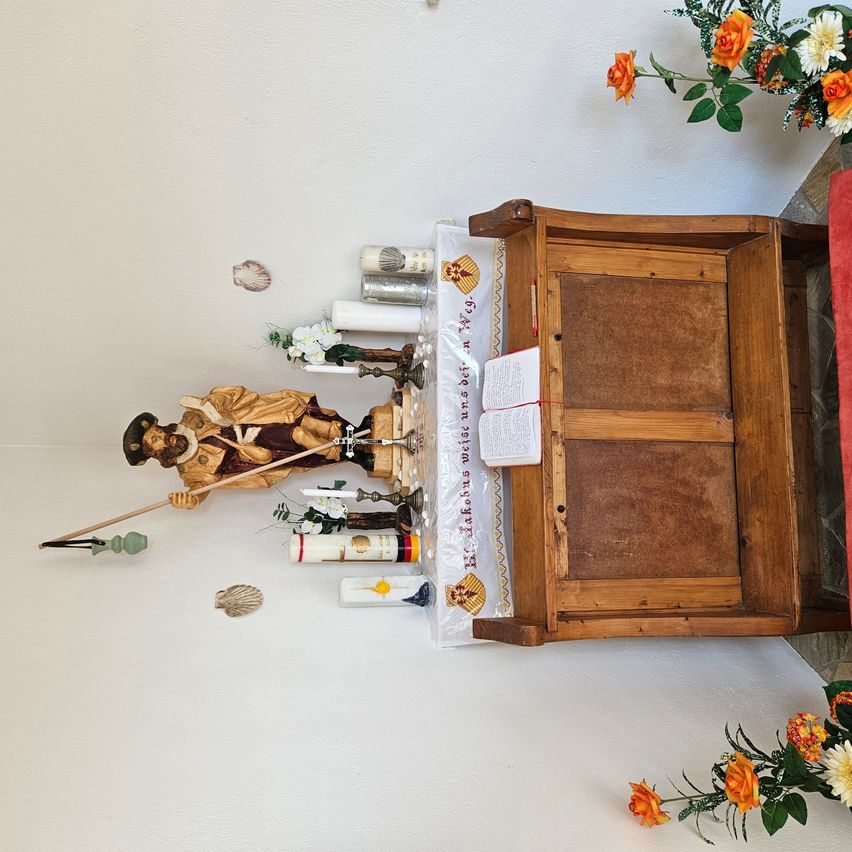Ein hölzerner Schrank mit einem Buch darauf steht an einer weißen Wand. Darauf befindet sich eine Statue eines Mannes mit einer Angelrute. Die Wand ist mit Blumen und Muscheln geschmückt.