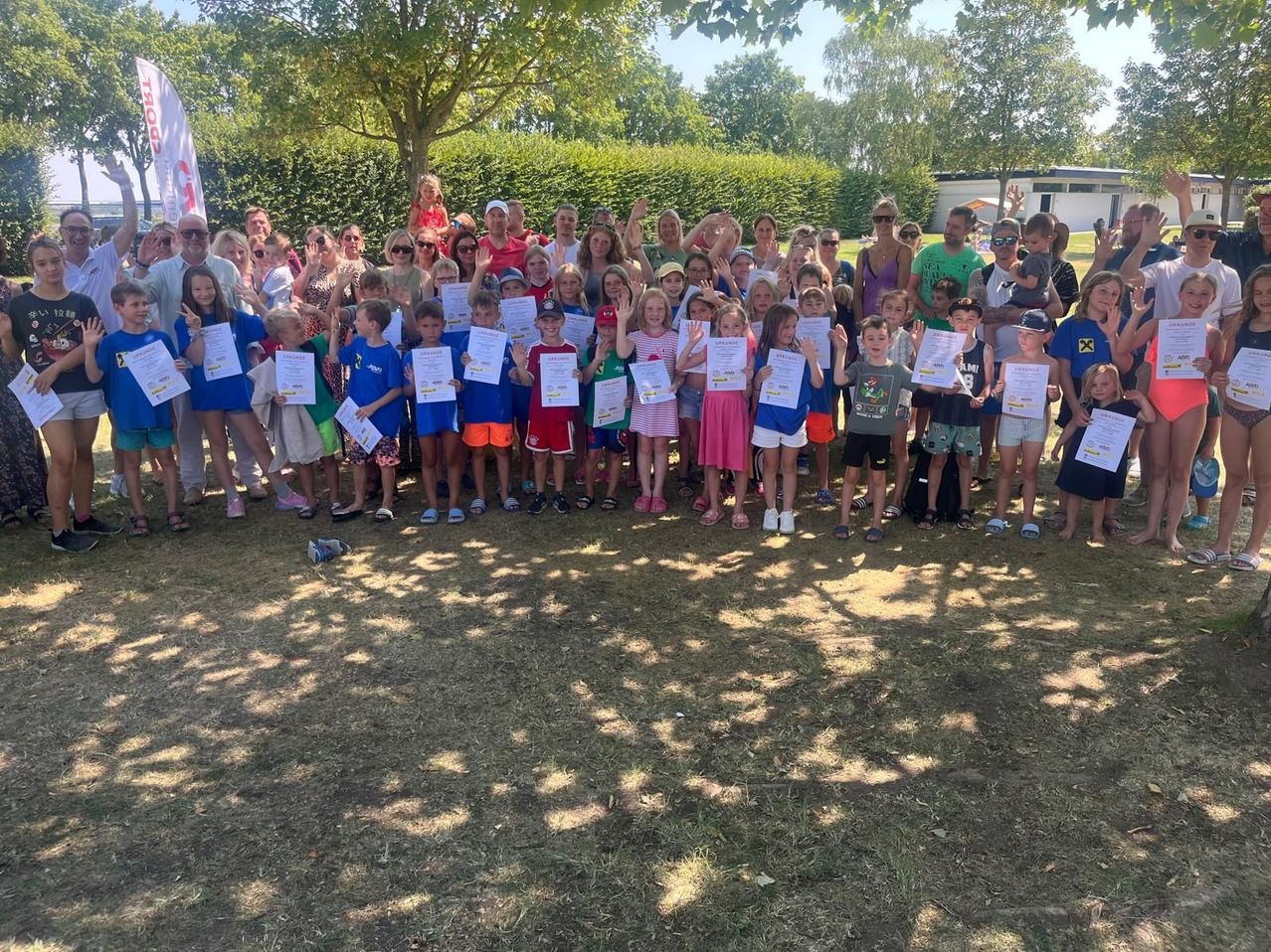 Eine Gruppe von Kindern und Erwachsenen versammelt sich in einem Park, hält Urkunden und posiert für ein Foto. Die Kinder tragen passende blaue Shirts, während Erwachsene verschiedene Outfits tragen. Bäume und ein Gebäude sind im Hintergrund zu sehen.