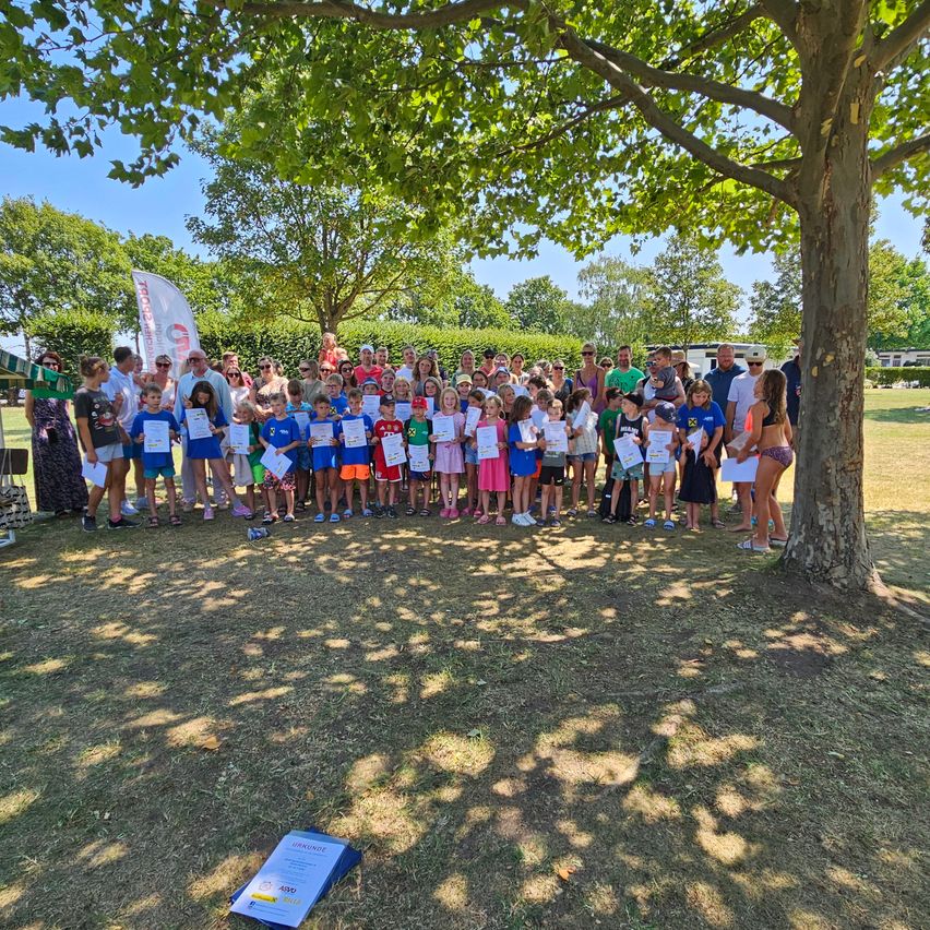 Eine Gruppe von Kindern und Erwachsenen steht unter einem Baum, hält Urkunden in der Hand, mit einer Flagge im Hintergrund.