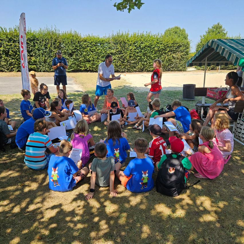 Eine Gruppe von Kindern und Erwachsenen sitzt im Kreis auf dem Gras, mit einem Mann und einer Frau, die vor ihnen stehen. Die Kinder halten Papiere in den Händen und scheinen aufmerksam zuzuhören.