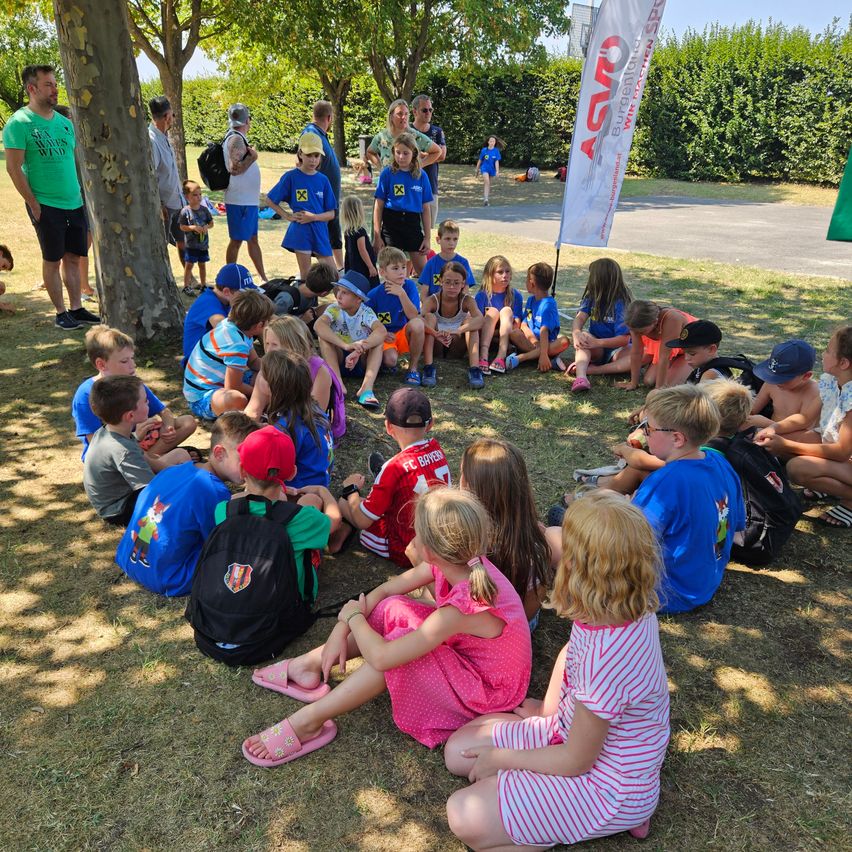Eine Gruppe von Kindern und Erwachsenen sitzt in einem Kreis auf einem Rasenfeld, einige tragen blaue Shirts mit einem Logo, während andere in der Nähe stehen oder sitzen. Bäume und ein Banner sind im Hintergrund sichtbar.