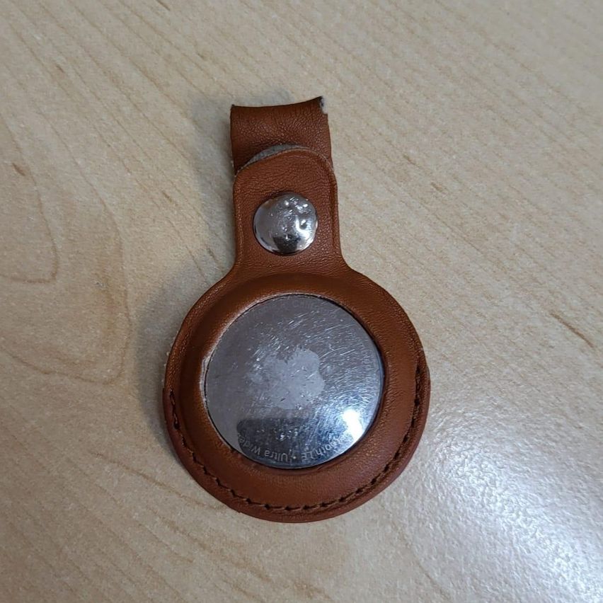Ein brauner Leder-Apple AirTag-Schlüsselanhänger mit einer silbernen runden Abdeckung und einem silbernen Knopf an der Seite, auf einer Holzoberfläche platziert.
