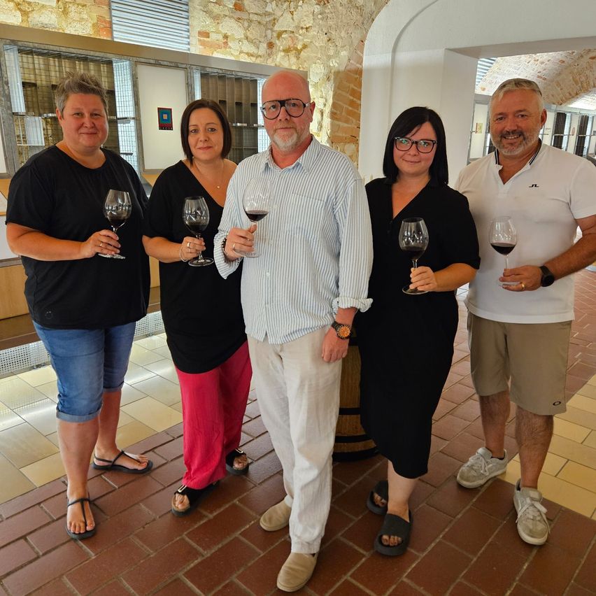 Fünf Personen, drei Frauen und zwei Männer, stehen zusammen in einem Raum, jeder hält ein Weinglas mit rotem Wein gefüllt, lächeln für ein Foto.