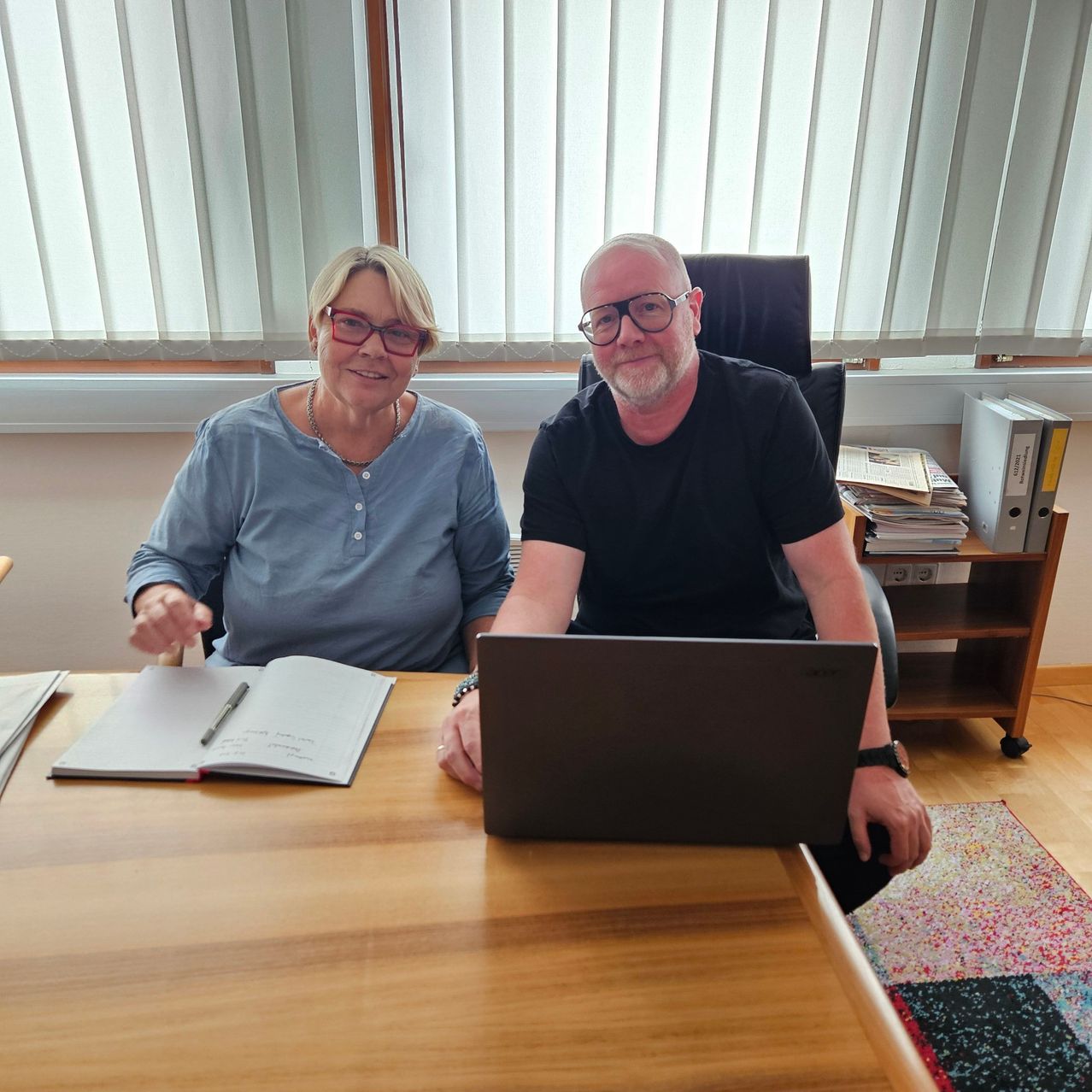 Ein Mann und eine Frau sitzen lächelnd für ein Foto am Schreibtisch. Die Frau trägt eine Brille und eine Halskette, und der Mann trägt eine Brille und eine Uhr. Sie haben einen Laptop und ein Notizbuch vor sich. Dahinter steht ein Regal mit Ordnern und Büchern.