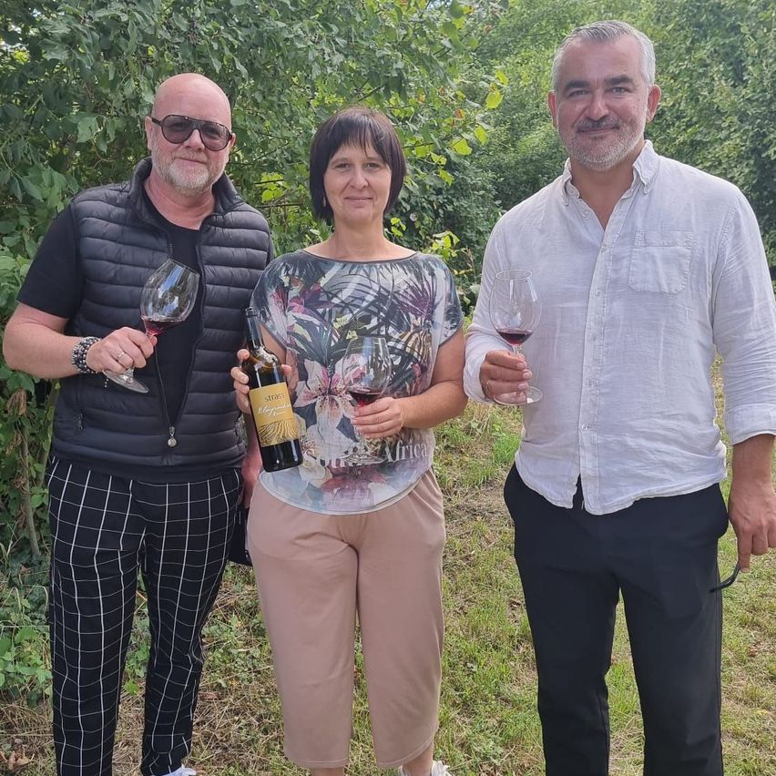 Drei Personen stehen in einem Weinberg, jede hält ein Glas Wein und eine Flasche. Der Mann links trägt eine schwarze Weste, die Frau in der Mitte ein Blumenoberteil und der Mann rechts ein weißes Hemd. Sie alle lächeln und schauen in die Kamera.