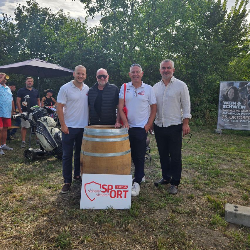 Vier Männer stehen um einen Holzfass mit einem Schild, das 'SP SPORT sicherer sicherer' besagt. Sie lächeln und scheinen für ein Foto zu posieren. Hinter ihnen stehen Bäume und ein Golfwagen.