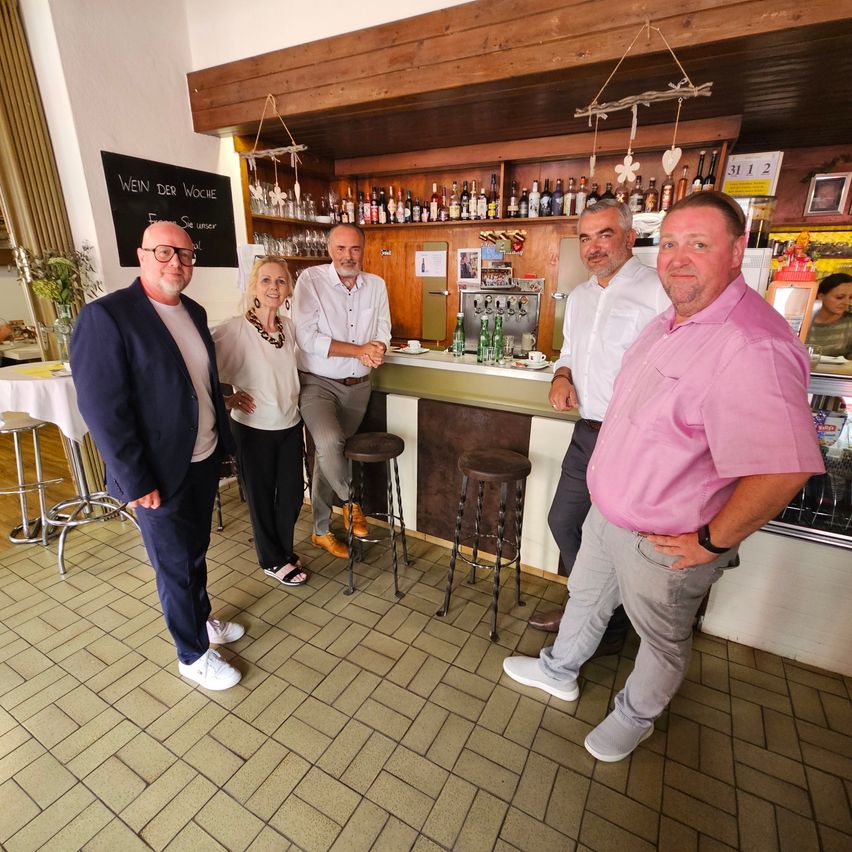 Fünf Menschen stehen in einer Bar, lächeln und posieren für ein Foto. Hinter ihnen befindet sich eine Theke mit Flaschen und einer Tafel.