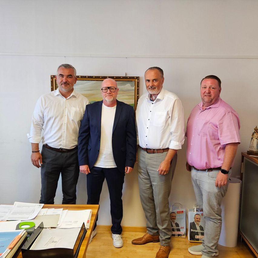 Vier Männer stehen in einem Büro und lächeln für ein Foto. Sie tragen verschiedenfarbige Hemden und Hosen, einer trägt eine Brille. Hinter ihnen hängt ein Gemälde an der Wand, und ein Schreibtisch mit Papieren und einer Tasche ist zu sehen.