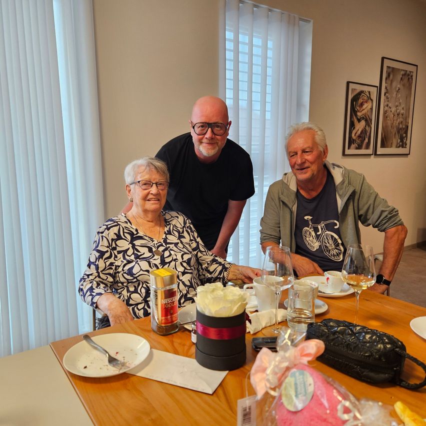Drei Personen sitzen an einem Esstisch und lächeln für ein Foto. Eine Frau in der Mitte hat einen Teller mit einer Gabel, und ein Mann hinter ihr hat eine Kaffeetasse. Verschiedene Gegenstände wie Weingläser, eine Handtasche und ein kleines Geschenk sind auf dem Tisch.