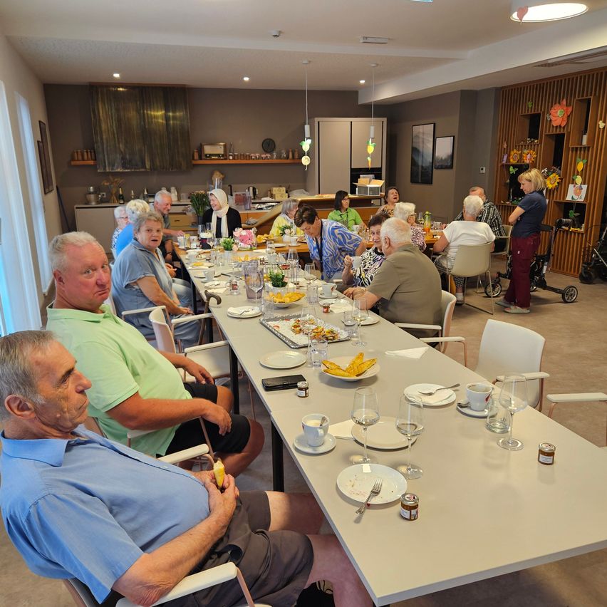 Eine Gruppe älterer Menschen sitzt um einen langen Tisch in einem Esszimmer und genießt ihr Essen. Der Tisch ist mit Tellern, Gläsern und verschiedenen Speisen gedeckt. Der Raum hat ein modernes Design mit Holzakzenten und einer hellen Atmosphäre.