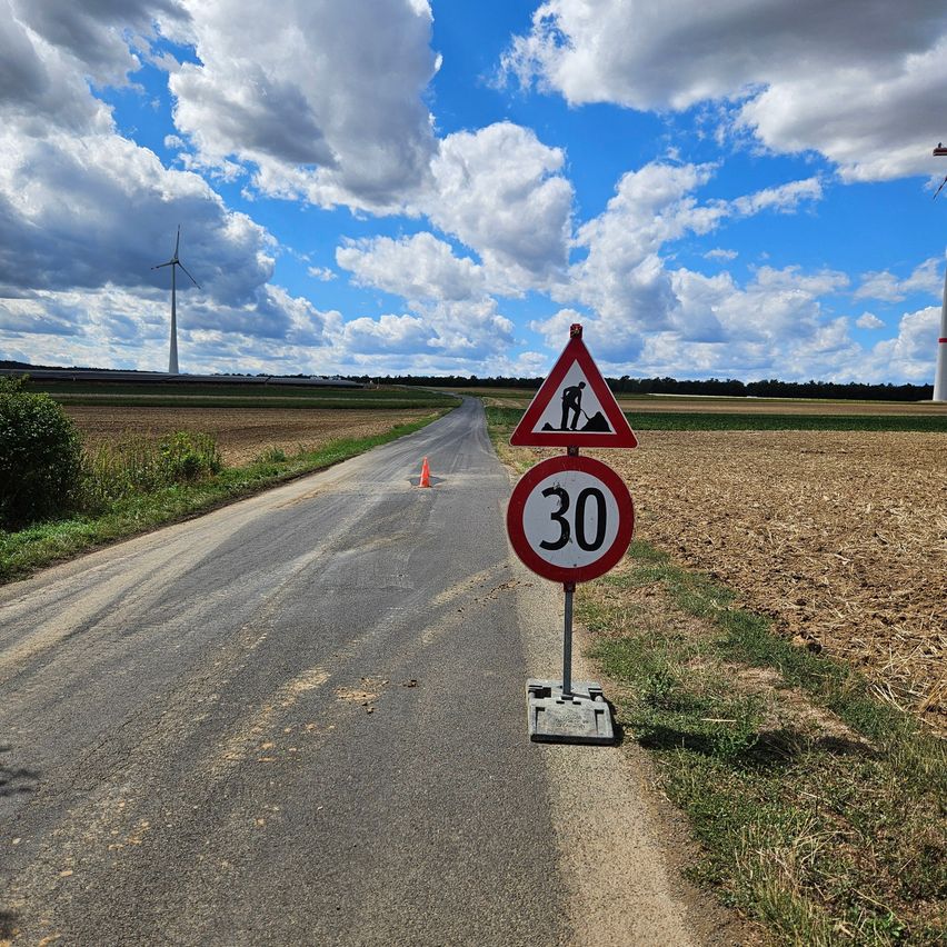Eine ländliche Straße mit einem Geschwindigkeitsbegrenzungsschild von 30 km/h, einem Baustellenwarnschild und einem Windrad in der Ferne unter einem bewölkten Himmel.