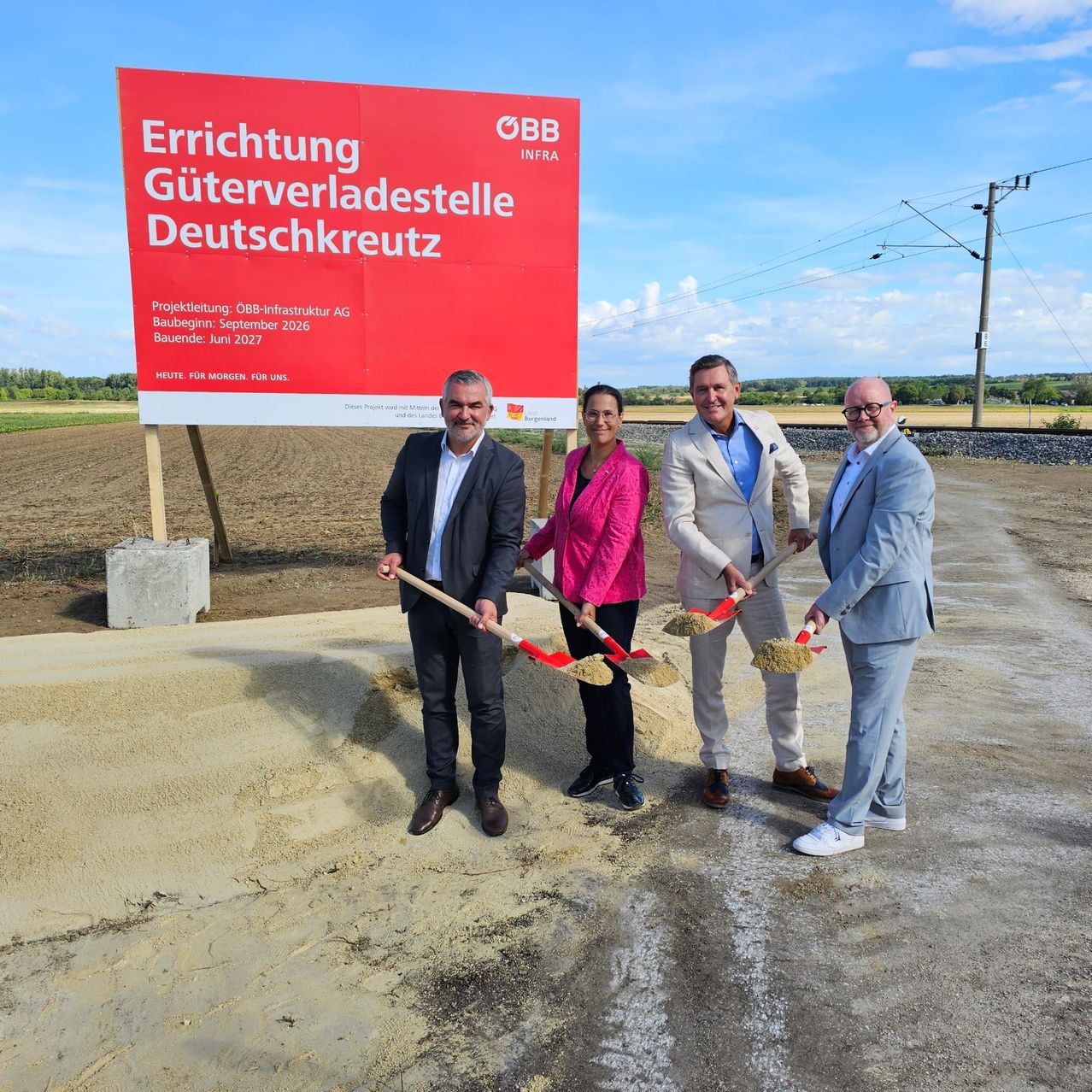 Four individuals in formal attire stand in front of a large red sign, holding shovels and posing for a photo. The sign reads 'Errichtung Güterverladestelle Deutschkreuz'. They are on a dirt road with a field and power lines in the background.
