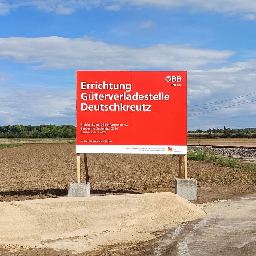 A red sign in a field indicates the construction of a goods transfer station. It reads 'Errichtung Güterverladestelle Deutschkreuz'. The sign is mounted on a wooden post and surrounded by a dirt field.