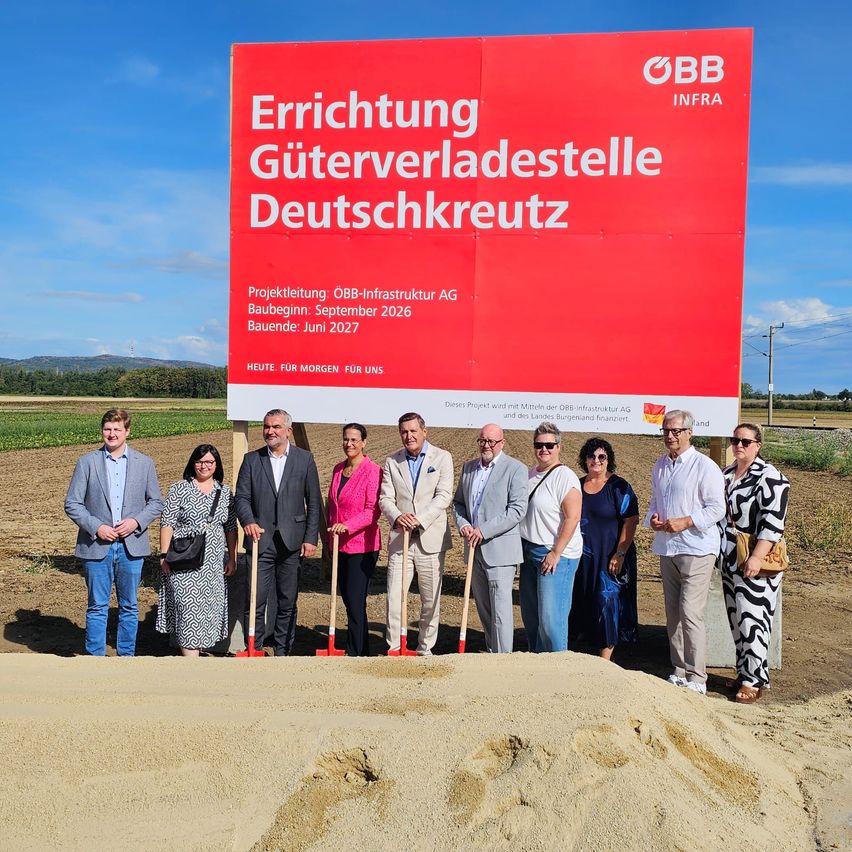 A group of people stand in front of a large red sign that reads 'Errichtung Guterverladestelle Deutschkreuz'. They are holding shovels and appear to be at a groundbreaking ceremony.