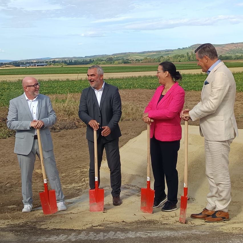 Four people in suits and glasses stand in a field, holding shovels. They appear to be starting a project.