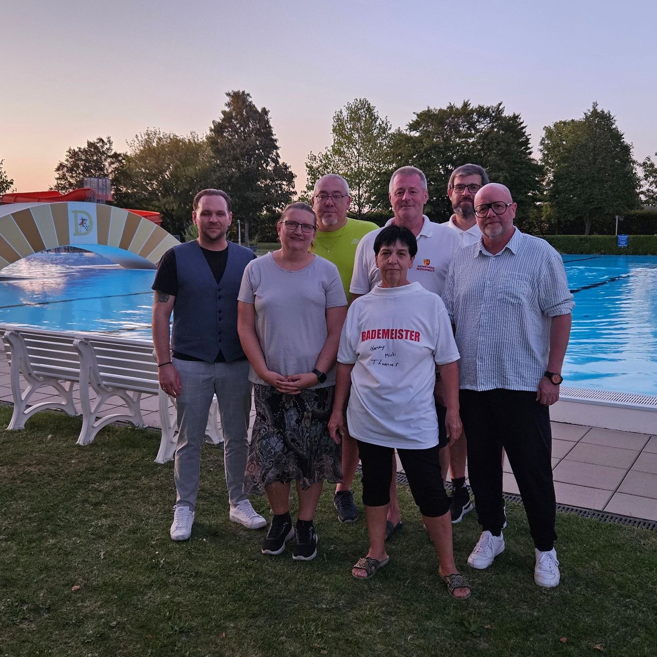 A group of people stands in front of a swimming pool, smiling for a photo. The pool has a bridge and slides in the background.