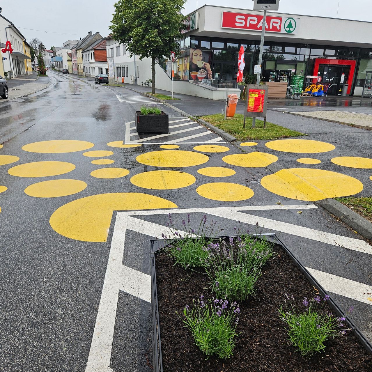 A wet road with yellow circles. A flowerbed and a sign are in the foreground. A SPAR supermarket is in the background.