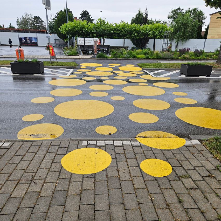 A wet road with yellow polka dots painted on it. Trees, plants, and a yellow building are visible in the background.