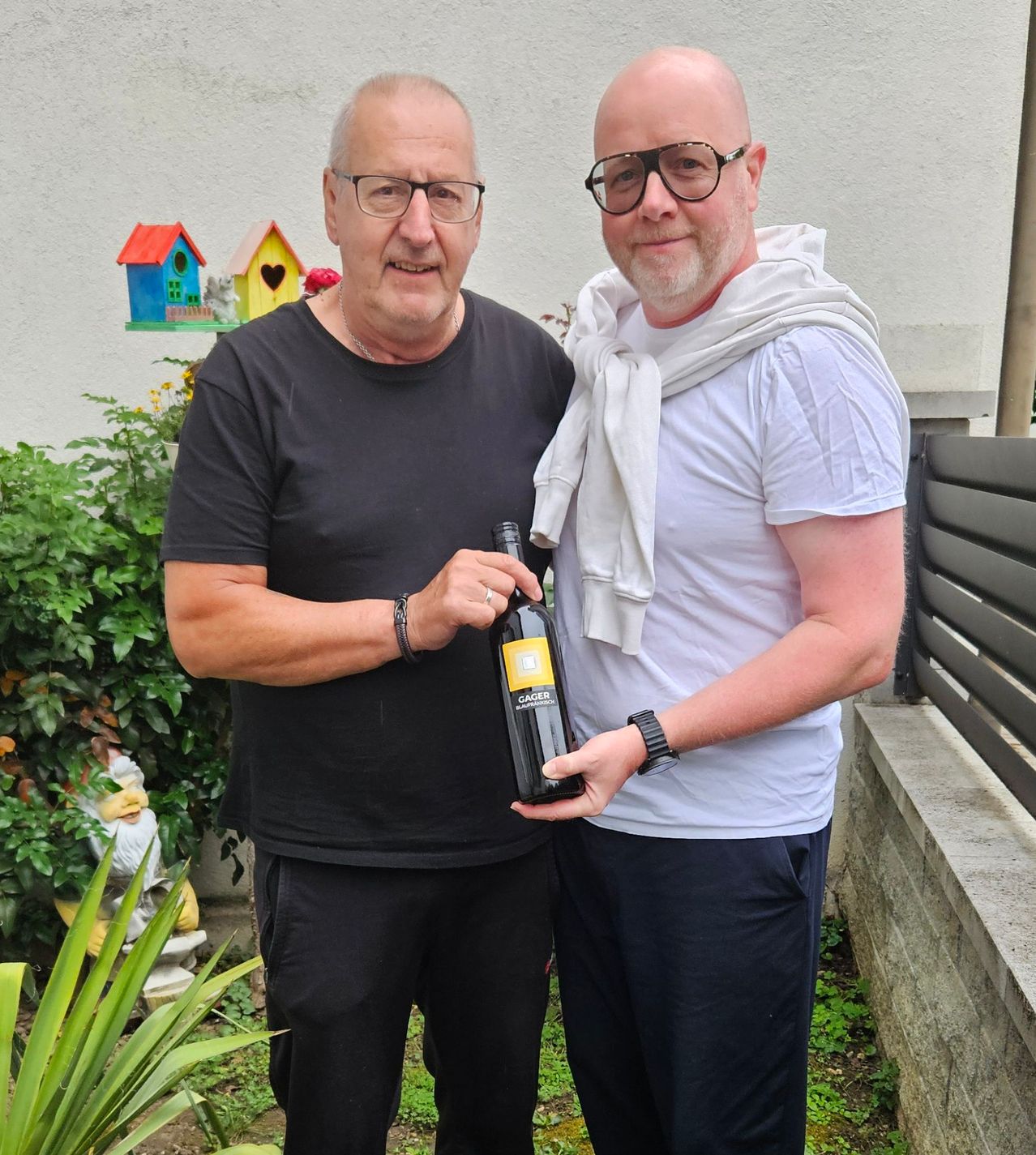 Two men stand in a garden, one holding a bottle of wine. The man on the left wears a black shirt and glasses, the other a white shirt and scarf. They stand in front of a house with a colorful birdhouse on the wall.