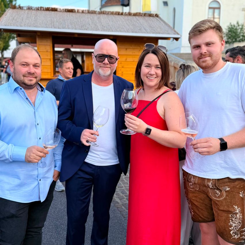 Four people stand in front of a wooden building, holding wine glasses. The man on the left wears a light blue shirt, the man in the middle a navy suit, the woman in a red dress, and the man on the right a white shirt and brown shorts.