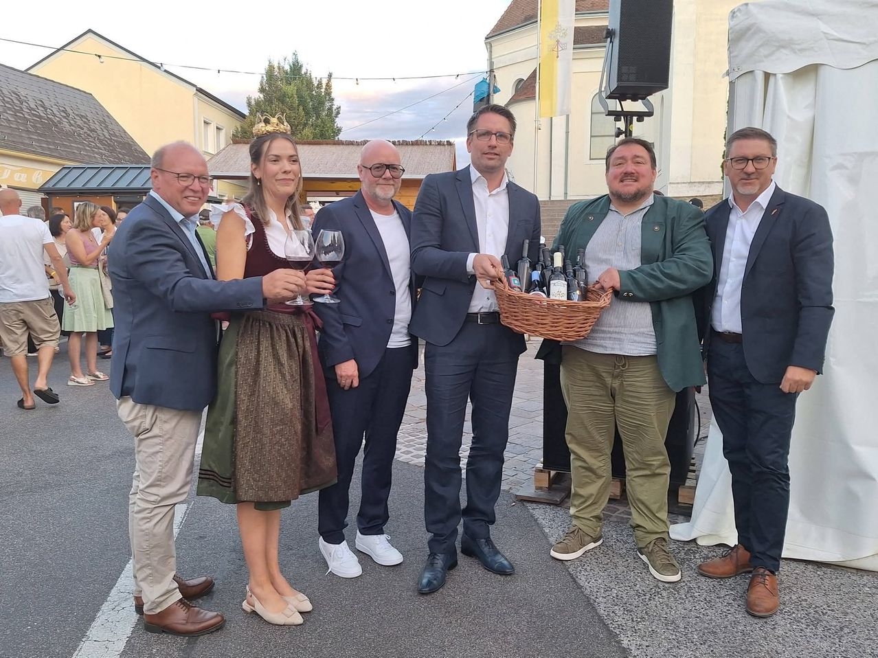 Six men and a woman stand in a group, smiling for a photo. The woman wears a crown and holds a wine glass. One man holds a basket of wine bottles. They are in front of a building with a flag.