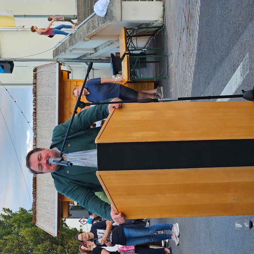 A man speaks into a microphone at a podium, with people standing behind him and others on a balcony above.
