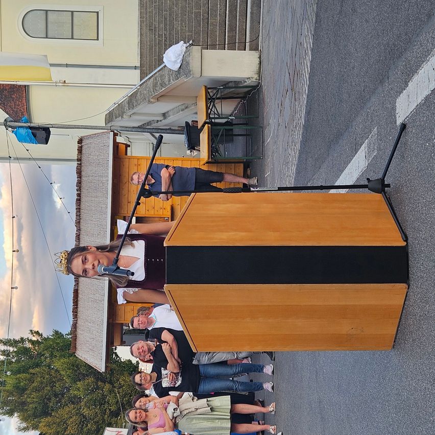 A girl wearing a crown is speaking into a microphone on a stage. Behind her, a group of people are standing and watching. The setting appears to be an outdoor event with a building and stairs in the background.