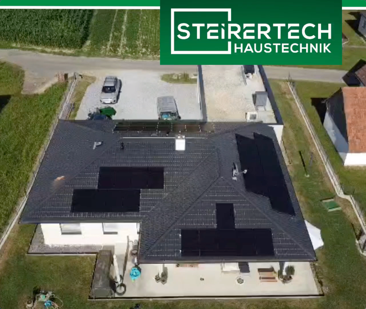 Luftaufnahme eines Hauses mit Solarpanelen auf dem Dach. Das Haus befindet sich in einer grünen Landschaft mit einem Parkplatz und Bäumen.