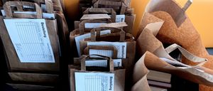 Ein Stapel brauner Papiertüten mit weißen Bestellformularen im Inneren. Jeder Beutel hat ein Etikett mit dem Wort 'Bestellung' und einer Checkliste. Einige Beutel enthalten Bücher.