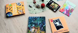 Bücher, Zeitschriften und ein kleiner Korb mit Gegenständen liegen auf einem Holzboden. Ein Buch trägt den Titel 'Peter Pan'.
