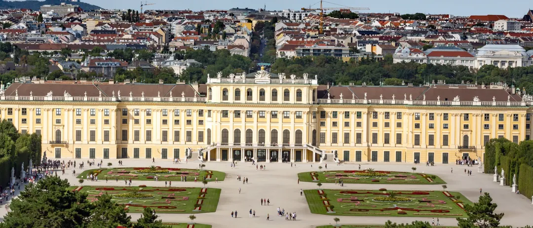 Ein Luftbild des Schönbrunner Schlosses mit seinen weitläufigen Gärten und der Stadtsilhouette im Hintergrund.