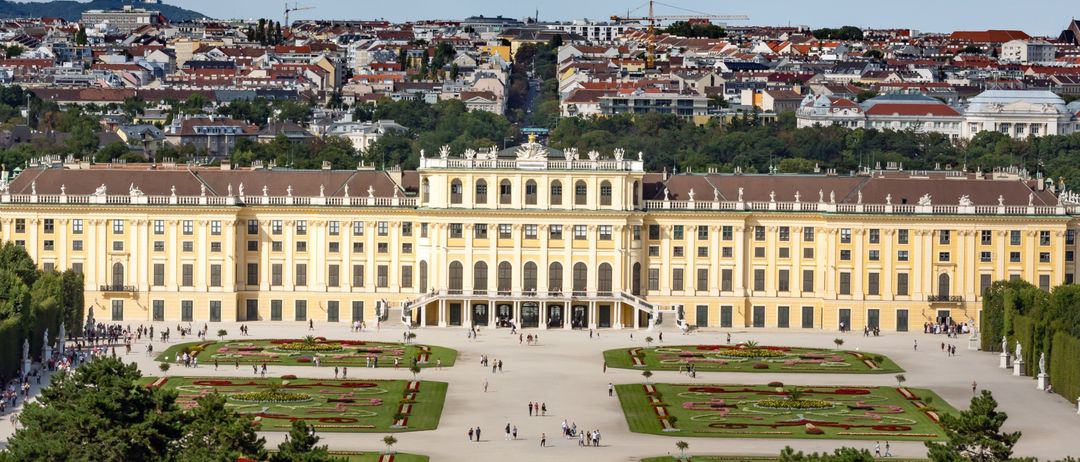 Ein Luftbild des Schönbrunner Schlosses mit seinen weitläufigen Gärten und der Stadtsilhouette im Hintergrund.
