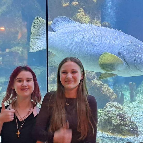 Zwei junge Frauen stehen vor einem großen Aquarium, eine gibt einen Daumen hoch. Dahinter schwimmt ein großer Fisch in der Nähe des Glases.