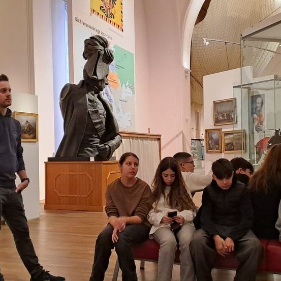 In einem Museum sitzen eine Gruppe von Menschen auf einer Bank, mit einem Mann in der Nähe einer Statue einer historischen Figur. Mehrere Gemälde schmücken die Wände, und ein Glasvitrine enthält verschiedene Gegenstände.