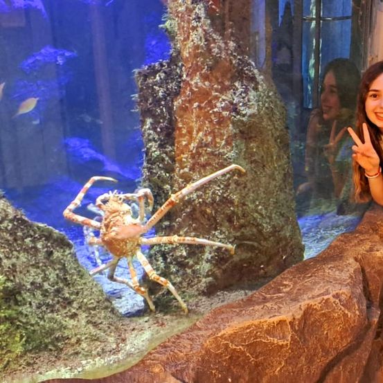 Eine lächelnde Frau macht ein Friedenszeichen neben einem Krabben in einem Aquarium mit blauem Wasser und Fischen.