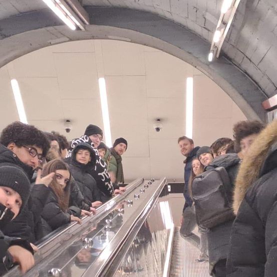 Eine Gruppe von Menschen steht auf einer Rolltreppe in einer U-Bahn-Station. Sie tragen Winterkleidung. Einige halten sich am Handlauf fest. Die Station hat gewölbte Decken und weiße Wände mit angebrachten Leuchten.