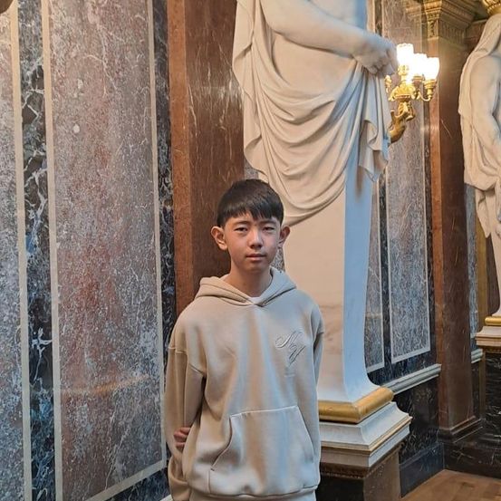 Ein junger Junge steht neben einer weißen Marmorstatue in einem prächtigen Raum. Der Junge trägt eine beigefarbene Kapuzenjacke.