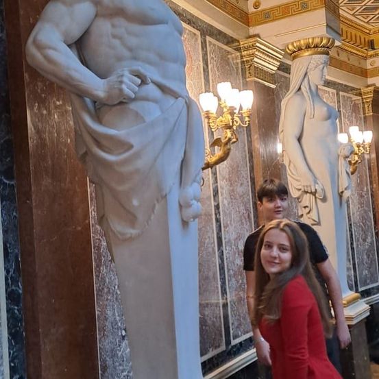 Eine Statue eines muskulösen Mannes und zwei Statuen von Frauen in einem Flur mit kunstvollen Marmorwänden. Ein Mädchen in einem roten Kleid und zwei Jungen stehen in der Nähe.