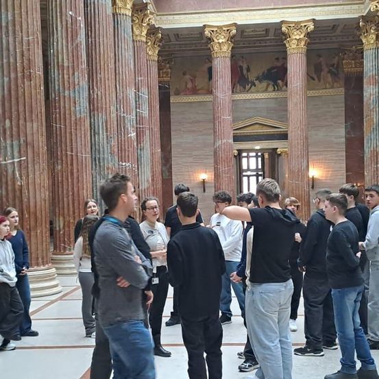 Eine Gruppe von Menschen, einige mit Brille und Turnschuhen, versammelt sich in einem geräumigen Saal mit Marmorsäulen. Einige lauschen einem Redner.