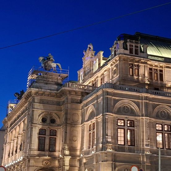 Ein Nachtblick auf ein großes Gebäude mit einer Statue eines Mannes auf einem Pferd oben darauf. Das Gebäude hat Bogenfenster, kunstvolle Details und wird von Lichtern beleuchtet.