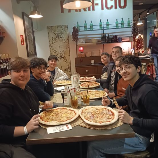 Eine Gruppe junger Männer sitzt an einem Tisch in einem Restaurant, lächelt und posiert für ein Foto. Es gibt mehrere Pizzen, Getränke und Gewürze auf dem Tisch. Dahinter steht ein Regal mit Flaschen und ein Schild an der Wand.