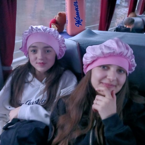 Zwei junge Mädchen mit rosa Kopftüchern sitzen lächelnd in einem Bus. Eines der Mädchen hat eine Tasche. Ein Plüschtier und eine Flasche sind im Hintergrund zu sehen.