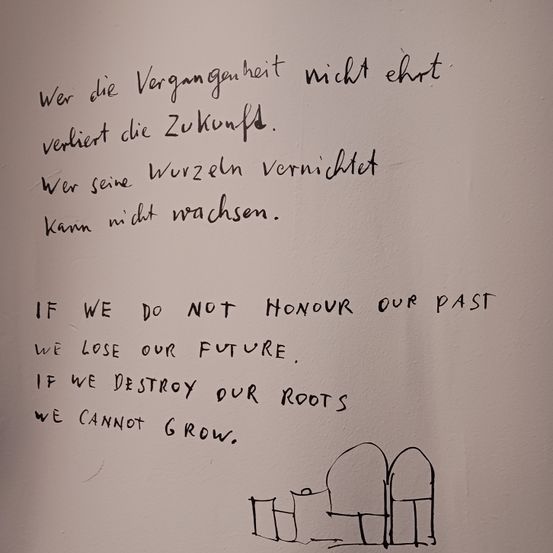 Ein handgeschriebener Zettel auf einer weißen Wand lautet: Wenn wir unsere Vergangenheit nicht ehren, verlieren wir unsere Zukunft. Wenn wir unsere Wurzeln zerstören, können wir nicht wachsen. Darunter ist eine Zeichnung von zwei Grabsteinen.