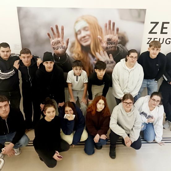 Eine Gruppe von Teenagern posiert für ein Foto vor einem großen Poster, wobei einige Hände halten und andere auf ihre Handys schauen. Sie tragen Winterkleidung. Das Poster zeigt eine Frau mit den Worten 'Unsere Zukunft' auf ihren Händen. Der Hintergrund ist unscharf.