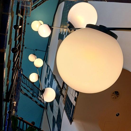 Ein Blick von unten zeigt eine Reihe kugelförmiger Lampen, die von der Decke hängen, jede mit einem schwarzen Rahmen und einer weißen Kugel. Die Lampen sind beleuchtet und werfen ein sanftes Licht. Der Hintergrund umfasst ein kariertes Muster an der Wand.